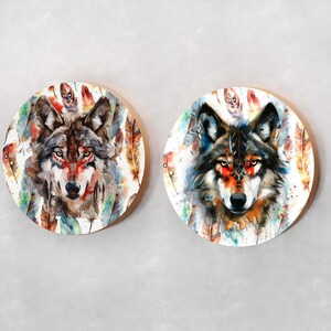 May include: Two round wooden wall decor pieces with watercolor paintings of wolf faces surrounded by colorful feathers. The wolf on the left has brown fur and the wolf on the right has gray fur.