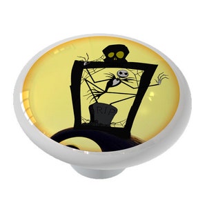 May include: A ceramic knob with a yellow background and a black silhouette of Jack Skellington from The Nightmare Before Christmas. He is standing in a window with a spiderweb and a tombstone that reads "RIP".