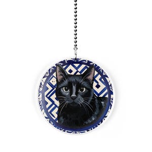 May include: A round, white ceiling fan pull featuring a black cat portrait. The cat has white whiskers and is centered on a blue and white geometric patterned background. A silver chain hangs from the top.
