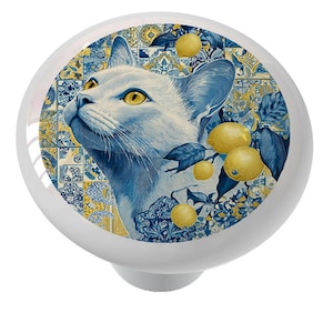 May include: White ceramic cabinet knob featuring a detailed illustration of a white cat with yellow eyes, surrounded by lemons and blue and yellow floral patterns. The knob has a rounded shape with a white base.