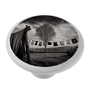 May include: A black and white photograph of a clothesline with various items of clothing hanging on it. The clothesline is in a rural setting with a tree in the background. The sky is cloudy and there is a crescent moon in the sky.