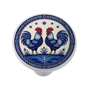 May include: White ceramic cabinet knob featuring a blue and white rooster design. Two roosters with red combs and blue tail feathers are centered on the knob. A blue floral border and accents complete the design. The knob is approximately 1.5 inches in diameter.