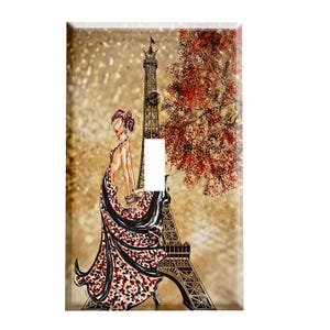 May include: A decorative light switch cover featuring a fashion illustration of a woman in a red and black patterned dress, with the Eiffel Tower and a tree with red leaves in the background. The cover has a gold glittery background.