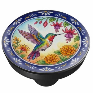 May include: A circular phone grip featuring a colorful illustration of a hummingbird in flight surrounded by orange marigolds and pink fuchsia flowers. The design is framed by a dark blue border with white daisy-like floral patterns.