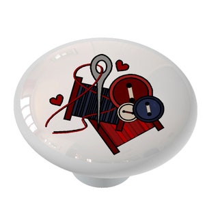 May include: A white ceramic knob with a red, black, and blue illustration of a spool of thread, a needle, and buttons. There are two red hearts in the design.
