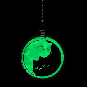 May include: A black and green glow-in-the-dark pendant featuring two cats in a yin yang design. The cats are facing each other with their tails intertwined.