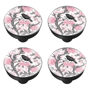 May include: Four black cabinet knobs with a floral design featuring pink flowers, gray branches, and a black bird. The knobs have a round, decorative top and a black base.