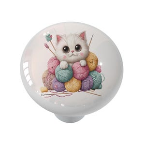 May include: White ceramic cabinet knob featuring a cartoon kitten nestled among colorful yarn balls. The kitten has large eyes and a pink nose. Knitting needles and a small knitted item are also visible. A decorative accent for furniture.
