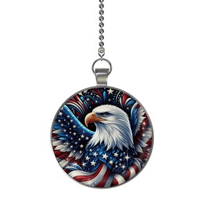 May include: A silver chain with a round pendant featuring a bald eagle with outstretched wings against a red, white, and blue American flag background. The eagle's head is turned to the side, and its eyes are looking to the right. The flag is stylized with stars and stripes, and there are fireworks in the background.
