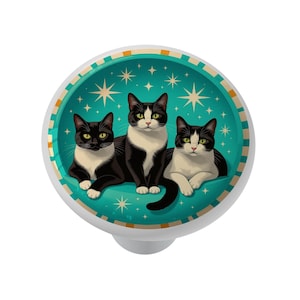 May include: A white cabinet knob with a teal background featuring three black and white cats. The cats are set against a backdrop of stars and a decorative border. The knob is a decorative element for furniture.