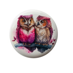 May include: Two watercolor owls perched on a branch with pink and brown feathers. The owl on the left has pink feathers and the owl on the right has brown feathers. The background is a watercolor wash of pink and blue.