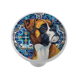 May include: A white cabinet knob featuring a Boxer dog portrait against a blue and white patterned background. The dog's fur is brown, tan, and white, with a black muzzle. The knob has a rounded shape and a silver-colored base.