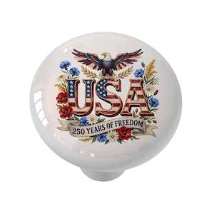 May include: A white ceramic cabinet knob featuring a patriotic design. The design includes the word "USA" in red, white, and blue, an eagle, and the text "250 YEARS OF FREEDOM" on a banner, surrounded by flowers.