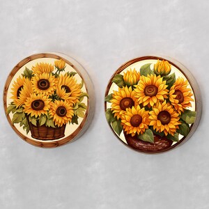 May include: Two round wooden coasters with a white background and a painted image of sunflowers in a basket. The sunflowers are yellow with brown centers and green leaves. The baskets are brown with a woven pattern.