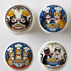 May include: Four white ceramic cabinet knobs with cat chef illustrations. Each knob features a different design of cats wearing chef hats, holding wine glasses, and serving pasta. The knobs have colorful floral borders.