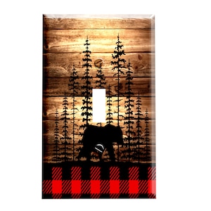 May include: A light switch cover with a rustic wood background and a silhouette of a bear walking through a forest of pine trees. The bottom of the cover features a red and black buffalo plaid pattern.