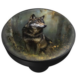 May include: A black round cabinet knob with a glass top featuring a detailed painting of a wolf in a forest setting. The wolf has brown and black fur, and the background includes trees and foliage.