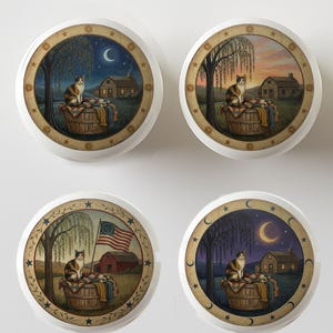 May include: Four round decorative cabinet knobs with a cat theme. Each knob features a different rural scene with a calico cat sitting on a basket. The backgrounds depict a night scene with a crescent moon, a sunset, a barn with an American flag, and a night scene.