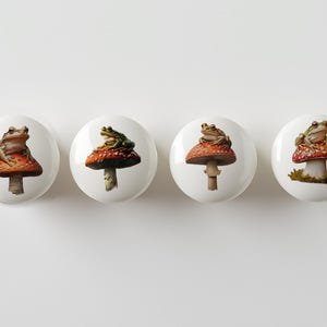 May include: Four ceramic knobs with a white background and a red and white mushroom with a frog on top. The frogs are different colors and patterns.