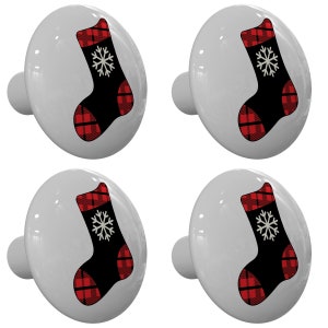 May include: Four white cabinet knobs with a Christmas stocking design. The black stocking has a red and black plaid cuff and toe, and a white snowflake in the center. The knobs are oval-shaped with a small, cylindrical stem.