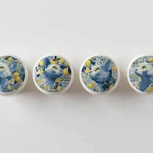 May include: Four round cabinet knobs with a cat and lemon design. The knobs feature a blue and yellow color scheme with a cat portrait surrounded by lemons and foliage. The knobs are white with a glossy finish.