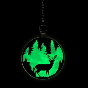 May include: A round, glowing pendant with a forest scene. The pendant features a black silhouette of a deer in front of a green, glowing forest and mountains. The pendant hangs from a silver chain against a black background.