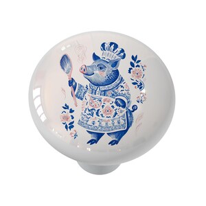 May include: White ceramic cabinet knob with a blue and pink illustration of a pig chef holding a spoon and a small bowl. The pig is wearing a chef's hat and apron with floral patterns. Decorative cabinet hardware.