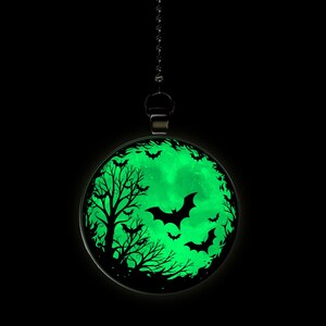 May include: A black metal pendant with a glow-in-the-dark moon and bats design. The moon is green and the bats are black. The pendant is hanging from a black chain.