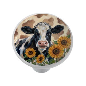 May include: White cabinet knob with a painted image of a black and white cow with sunflowers. The background features a brown and tan cowhide pattern. The knob is round and has a silver-colored base.