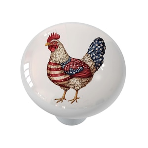 May include: A white ceramic cabinet knob featuring a detailed illustration of a rooster. The rooster's body is adorned with a design of the American flag, including red and white stripes and blue stars. The knob has a smooth, glossy finish.