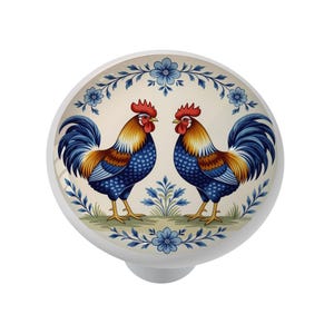 May include: White cabinet knob with a decorative design featuring two roosters facing each other. The roosters have blue, gold, and red plumage, set against a cream background with blue floral accents. A charming addition to any cabinet.