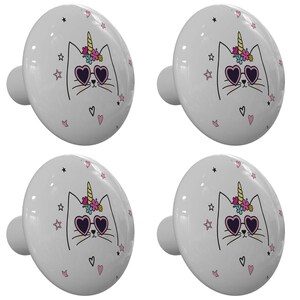 May include: Set of four white ceramic cabinet knobs with a unicorn cat design. The cat has pink heart-shaped sunglasses, a yellow horn, and a pink flower crown. The background is white with pink hearts and silver stars.