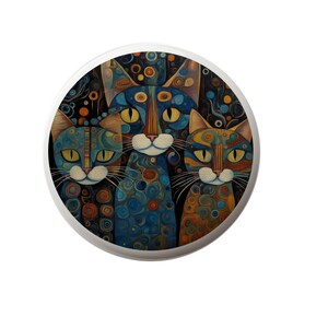 May include: A round decorative item featuring a colorful illustration of three stylized cats. The cats have blue, orange, and brown patterns with yellow eyes, set against a dark background with circular designs. The item has a white border.