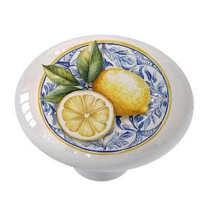 May include: A white ceramic knob with a blue and yellow design featuring lemons and leaves. The knob has a raised edge and a smooth surface.