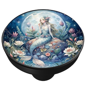 May include: A black cabinet knob featuring a watercolor illustration of a mermaid sitting on a rock in a moonlit scene. The mermaid has long blonde hair and a blue and white tail. The background includes flowers and water.