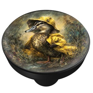 May include: A round, black pop-up phone grip featuring a detailed illustration of a duck wearing a yellow raincoat and hat. The duck is standing in tall grass, with a rainy background.