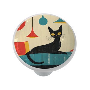 May include: A white cabinet knob with a retro illustration of a black cat with yellow eyes. The cat is lounging on a blue surface, with red and blue pendant lights in the background. The knob has a silver base.