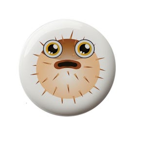 May include: A round button with a cartoon image of a brown and white pufferfish with big eyes and a surprised expression.