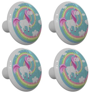 May include: Four white ceramic cabinet knobs with a unicorn design. Each knob features a white unicorn with a pink mane and tail standing on a rainbow with stars and clouds. The background is a light blue color.