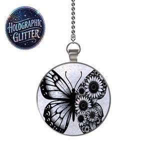 May include: A round ceiling fan pull chain ornament featuring a butterfly design. The left wing is a solid black butterfly pattern, while the right wing is composed of black sunflowers. The background is a light holographic glitter finish.