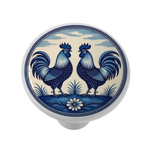 May include: White ceramic cabinet knob with a blue and white rooster design. The knob features two facing roosters, grass, and a floral element, all within a circular border. The design is reminiscent of classic blue and white pottery.