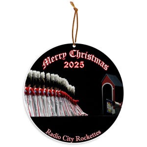May include: Round Christmas ornament with a black background featuring the Radio City Rockettes in red and white uniforms. The words "Merry Christmas 2025" are in red script. The words "Radio City Rockettes" are at the bottom.
