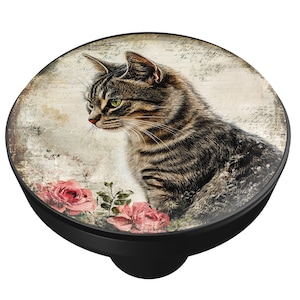 May include: A black phone grip with a round, printed top featuring a tabby cat with green eyes and pink roses. The cat is set against a vintage-style background with script. The grip has a black base.