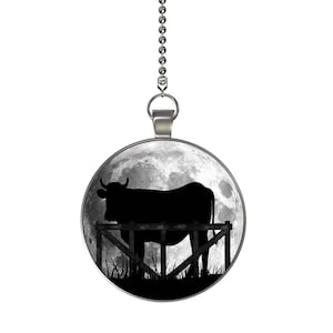 May include: A silver chain with a round pendant featuring a black silhouette of a cow standing in a field with a full moon in the background.