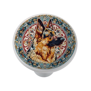May include: A white cabinet knob featuring a German Shepherd dog illustration. The dog is depicted in shades of brown, black, and tan, set against a decorative circular background with blue, red, and gold accents. The knob has a rounded top and a short, cylindrical base.