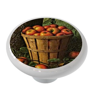 May include: A ceramic knob with a vintage illustration of a wooden basket overflowing with red apples. The basket is surrounded by green leaves and brown branches.