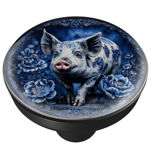 May include: A decorative bottle stopper featuring a detailed illustration of a pig with blue and white spots. The pig is set against a blue floral background. The stopper has a black base.