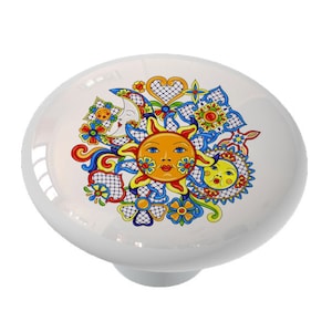 May include: A white ceramic knob with a colorful sun, moon, and floral design. The sun and moon have faces and are surrounded by flowers and hearts.