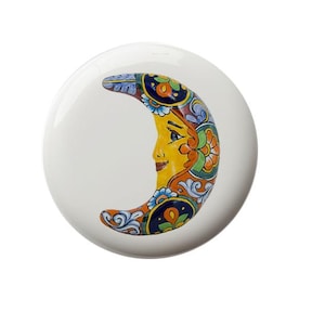 May include: A ceramic crescent moon wall decor with a colorful, hand-painted design. The moon features a face with blue eyes and a yellow background. The design includes orange, blue, green, and white floral patterns.