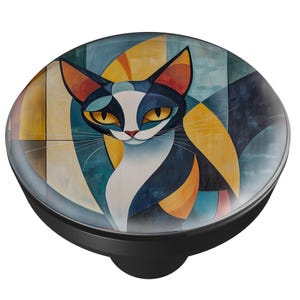 May include: A black cabinet knob with a colorful cat design. The cat has a white face, blue and black fur, and yellow eyes. The background features geometric shapes in yellow, blue, and orange.